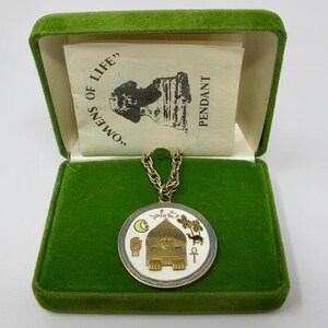 May include: A gold-colored pendant with a circular design, featuring an Egyptian-style structure, symbols, and text, displayed in a green velvet box. The pendant hangs from a chain. The box contains a paper with text and an illustration.