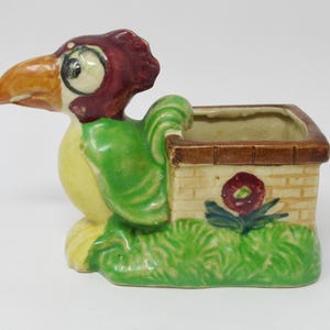 May include: A ceramic planter shaped like a cartoon bird with a green body, yellow belly, red head, and brown beak. The planter has a small rectangular pot on its back with a brown rim and a painted flower design.