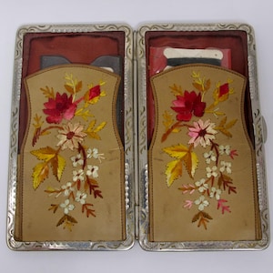 May include: A rectangular silver-toned metal case with an engraved border, opened to reveal two tan leather inserts. Each insert features an embroidered floral design in shades of red, pink, yellow, and white, with dark red fabric lining the interior.