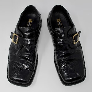 May include: A pair of black leather dress shoes with a buckle strap. The shoes have a textured, alligator-like pattern and a square toe.
