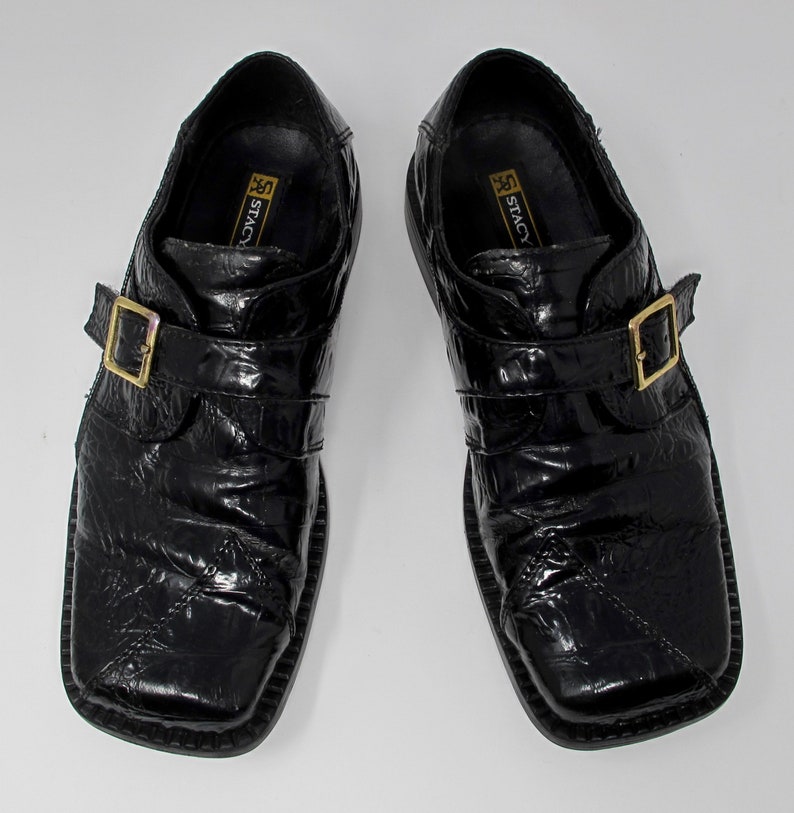 May include: A pair of black leather dress shoes with a buckle strap. The shoes have a textured, alligator-like pattern and a square toe.