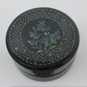 May include: A round, black lacquered box with a decorative lid. The lid features a floral design with iridescent accents and a geometric border. The box is likely used for jewelry or small keepsakes.