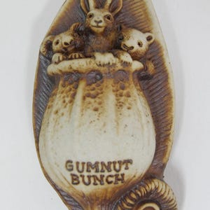May include: A ceramic Gumnut Bunch figurine. The figurine features a rabbit and two koalas emerging from a gum nut. The figurine is brown and beige with the words "Gumnut Bunch" at the bottom.