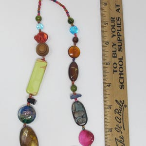 May include: A colorful necklace with a variety of beads, including wood, glass, and stone. The necklace is approximately 18 inches long and features a silver chain clasp. "Make It A Rule" and "To Buy Your School Supplies At Gamble" are printed on a wooden ruler in the background.