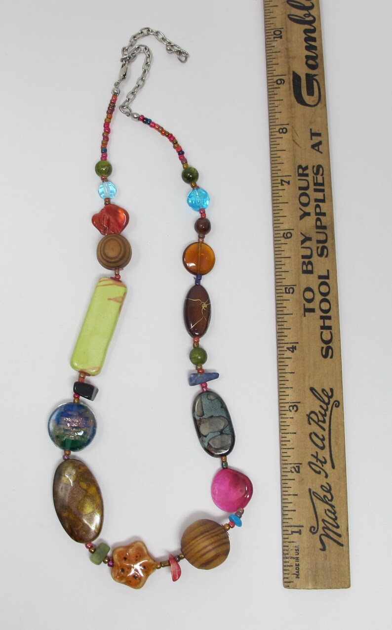 May include: A colorful necklace with a variety of beads, including wood, glass, and stone. The necklace is approximately 18 inches long and features a silver chain clasp. "Make It A Rule" and "To Buy Your School Supplies At Gamble" are printed on a wooden ruler in the background.