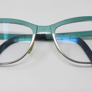 May include: A pair of eyeglasses with a teal-colored frame and silver rims. The temples are black, and the lenses are clear. The glasses are set against a white background.