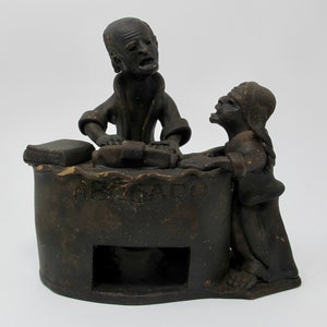 May include: A ceramic sculpture of two figures, one standing and one seated, with the word "ABOGADO" inscribed on the base. The figures are dark brown and appear to be engaged in a conversation.