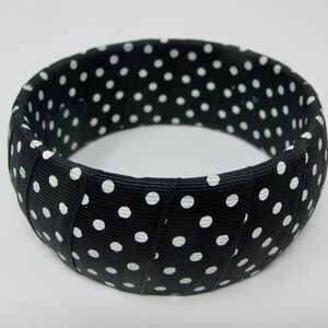 May include: Black and white polka dot fabric bangle bracelet. The bracelet is made of a thick, woven fabric with white polka dots on a black background.