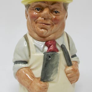 May include: A ceramic butcher figurine with a yellow hat, white apron, and a red tie. The figurine is holding a cleaver and a knife. The butcher has a smiling expression and is wearing a white shirt.