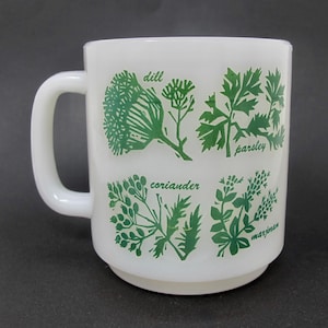 May include: A white milk glass mug featuring green botanical illustrations of herbs. The design includes four distinct plant graphics labeled with the text dill, parsley, coriander, and marjoram printed in a green script font on the side.