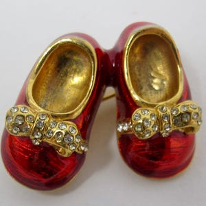 May include: A vintage brooch featuring a pair of red enamel baby shoes with gold-tone interiors and trim. Each shoe has a gold bow embellished with clear rhinestones. The brooch has a pin closure on the back.