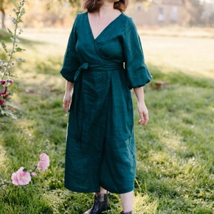 May include: A dark teal wrap dress with a V-neck and three-quarter sleeves. The dress is tied at the waist with a matching fabric belt. The model is wearing dark blue ankle boots with white polka dots. The background is a grassy field with a house in the distance.