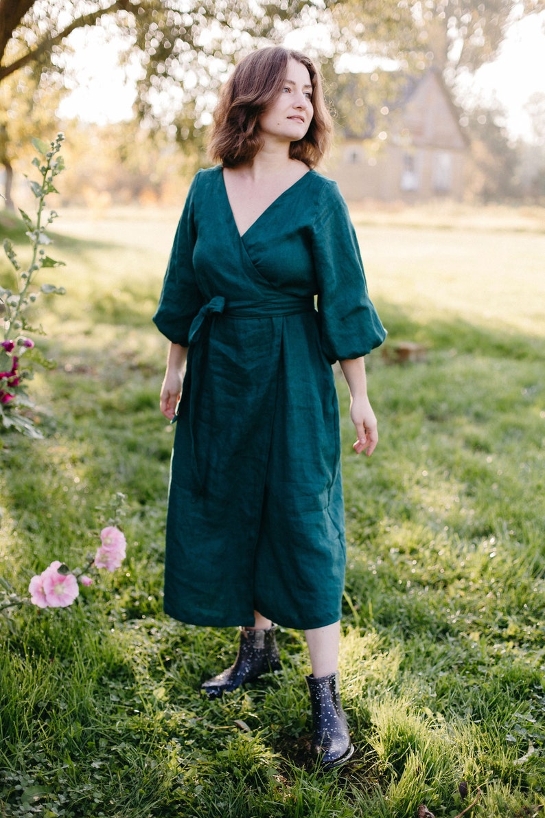 May include: A dark teal wrap dress with a V-neck and three-quarter sleeves. The dress is tied at the waist with a matching fabric belt. The model is wearing dark blue ankle boots with white polka dots. The background is a grassy field with a house in the distance.