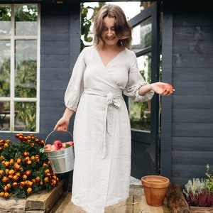 May include: A light gray wrap dress with long sleeves and a matching belt. A woman is holding a metal bucket filled with red apples and another apple in her other hand. The background includes a dark gray shed and a garden.