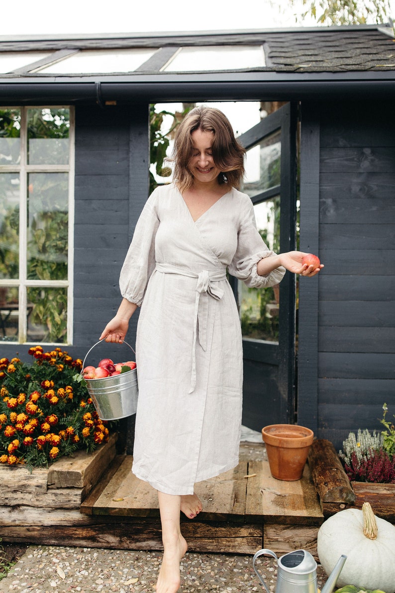 May include: A light gray wrap dress with long sleeves and a matching belt. A woman is holding a metal bucket filled with red apples and another apple in her other hand. The background includes a dark gray shed and a garden.