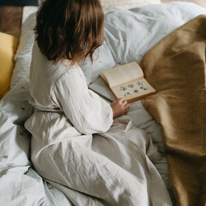 May include: A person wearing a light beige linen robe sits on a bed with a light blue patterned comforter and a tan blanket. The person is reading a book with illustrations. The room has a wooden cabinet and a woven basket.