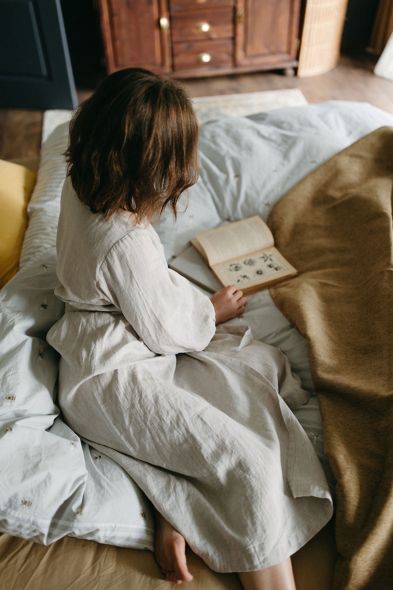 May include: A person wearing a light beige linen robe sits on a bed with a light blue patterned comforter and a tan blanket. The person is reading a book with illustrations. The room has a wooden cabinet and a woven basket.