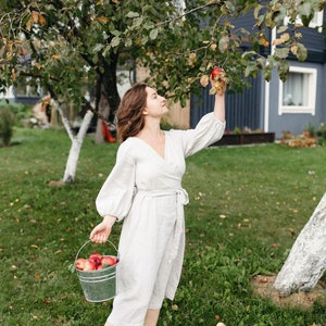 May include: A person in a light-colored linen wrap dress picks apples from a tree in a garden. A metal bucket filled with red apples is held in one hand. The scene is set in a grassy yard with a house in the background.