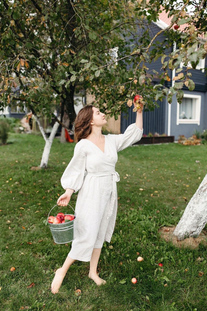 May include: A person in a light-colored linen wrap dress picks apples from a tree in a garden. A metal bucket filled with red apples is held in one hand. The scene is set in a grassy yard with a house in the background.