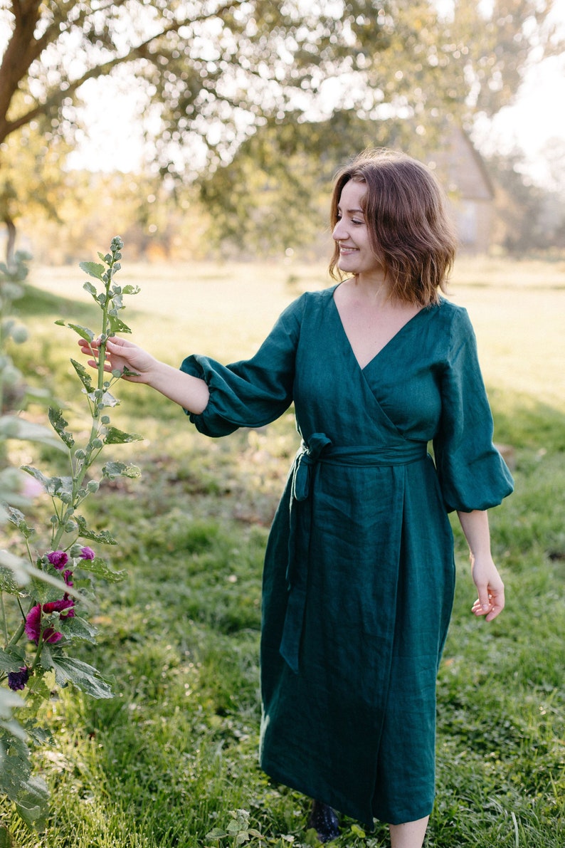 May include: A dark teal wrap dress with long, puffy sleeves and a matching waist tie. The dress has a V-neckline and falls to mid-calf length. The woman is standing in a grassy field, touching a tall plant with purple flowers.