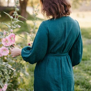 May include: A teal green linen dress with long, voluminous sleeves and a belted waist. The dress is knee-length and worn outdoors, with a person standing near pink hollyhock flowers. The background includes a house and trees.