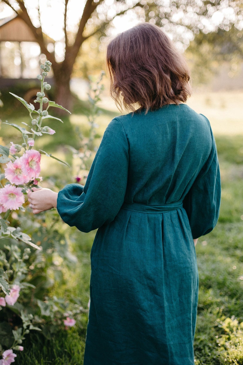 May include: A teal green linen dress with long, voluminous sleeves and a belted waist. The dress is knee-length and worn outdoors, with a person standing near pink hollyhock flowers. The background includes a house and trees.