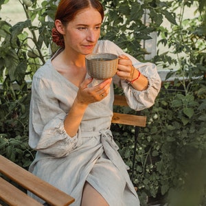May include: A woman in a light gray linen wrap dress sits on a wooden chair, holding a patterned ceramic mug. The scene is set in a greenhouse with lush green foliage. The dress has long sleeves and a tie waist.