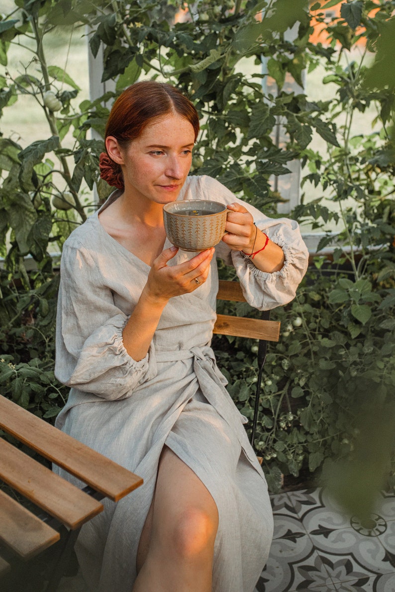 May include: A woman in a light gray linen wrap dress sits on a wooden chair, holding a patterned ceramic mug. The scene is set in a greenhouse with lush green foliage. The dress has long sleeves and a tie waist.