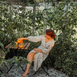 May include: A person in a light-colored wrap dress sits at a small wooden table in a greenhouse, pouring liquid from a glass container into a yellow mug. The greenhouse has a tiled floor and is filled with green plants.
