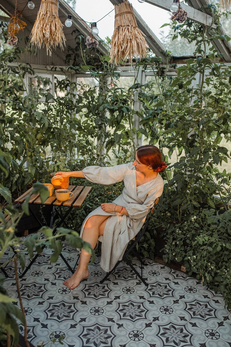 May include: A person in a light-colored wrap dress sits at a small wooden table in a greenhouse, pouring liquid from a glass container into a yellow mug. The greenhouse has a tiled floor and is filled with green plants.