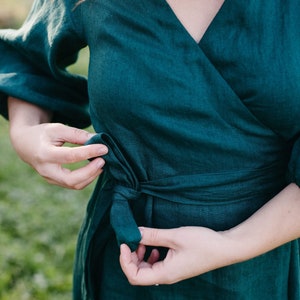 May include: A teal wrap dress made of linen. The dress features a V-neckline, long sleeves, and a tie closure at the waist. The fabric has a textured appearance. The dress is being tied by a person.
