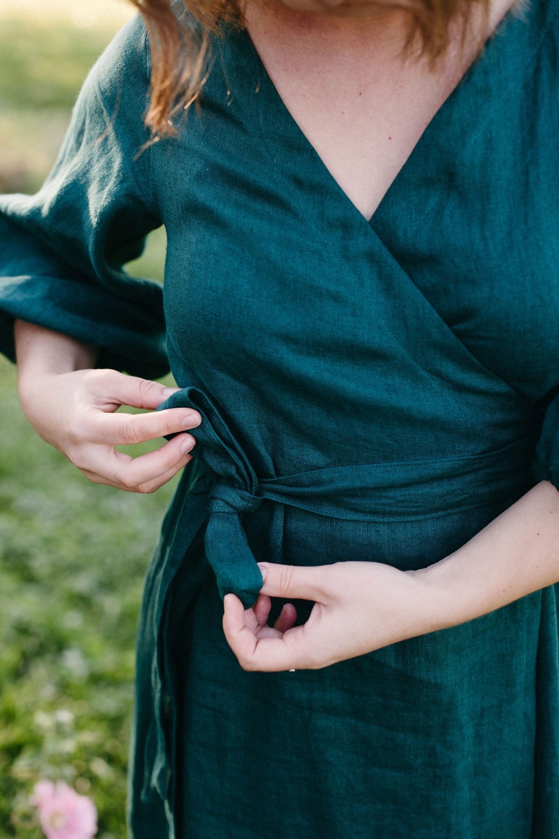 May include: A teal wrap dress made of linen. The dress features a V-neckline, long sleeves, and a tie closure at the waist. The fabric has a textured appearance. The dress is being tied by a person.