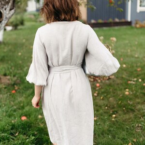 May include: A light beige linen dress with a wrap-around waist and elbow-length sleeves. The dress is knee-length and is being worn outdoors on a grassy lawn. The sleeves are puffy. The person is holding a silver tray.