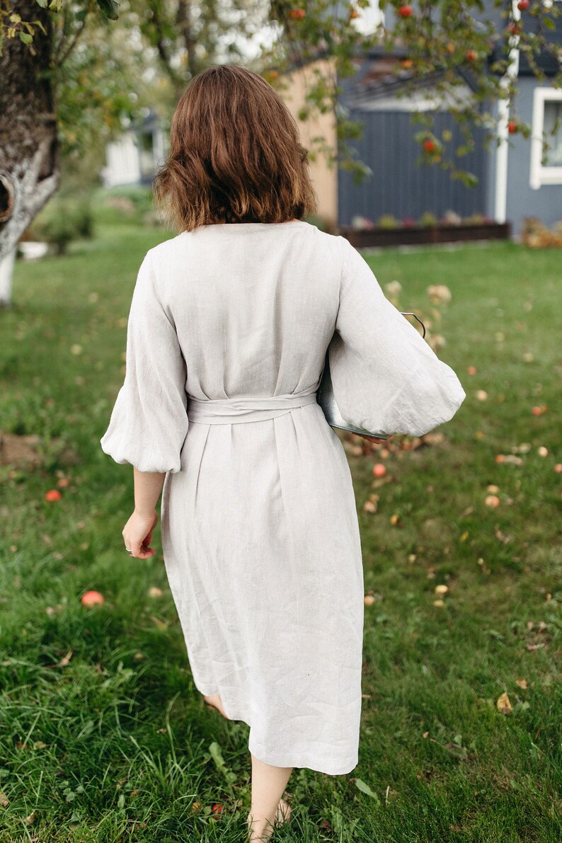 May include: A light beige linen dress with a wrap-around waist and elbow-length sleeves. The dress is knee-length and is being worn outdoors on a grassy lawn. The sleeves are puffy. The person is holding a silver tray.