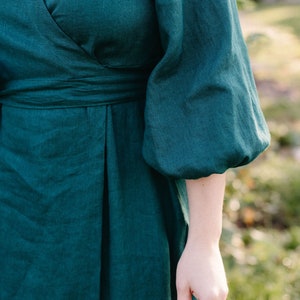 May include: Emerald green wrap dress with elbow-length sleeves. The dress features a V-neckline and a fitted waist. The fabric appears to be linen or a similar natural material, with a subtle texture. The dress is a solid color, with no visible patterns.