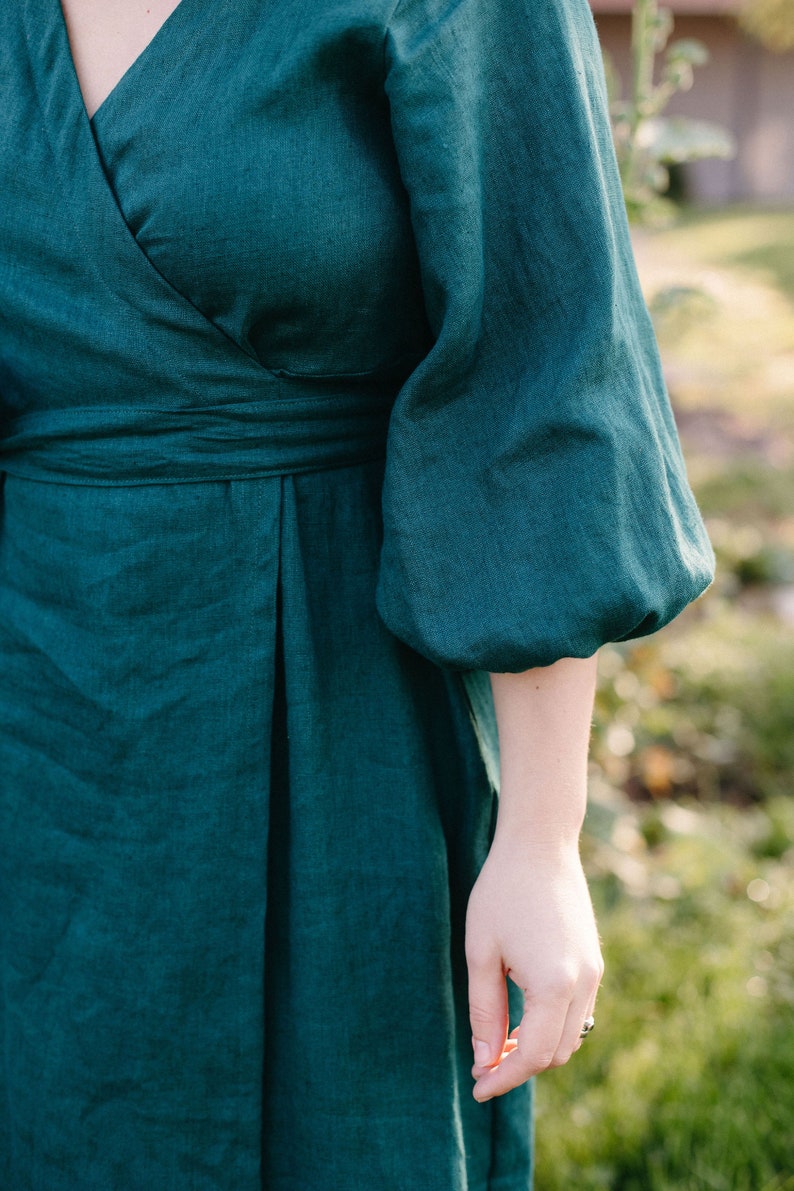 May include: Emerald green wrap dress with elbow-length sleeves. The dress features a V-neckline and a fitted waist. The fabric appears to be linen or a similar natural material, with a subtle texture. The dress is a solid color, with no visible patterns.