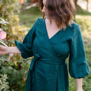 May include: A teal wrap dress with a V-neckline and long, puffy sleeves. The dress is tied at the waist with a matching fabric belt. The model is standing outside near pink flowers and greenery.