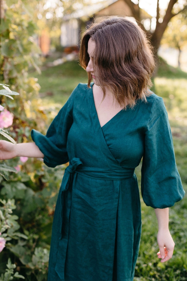 May include: A teal wrap dress with a V-neckline and long, puffy sleeves. The dress is tied at the waist with a matching fabric belt. The model is standing outside near pink flowers and greenery.