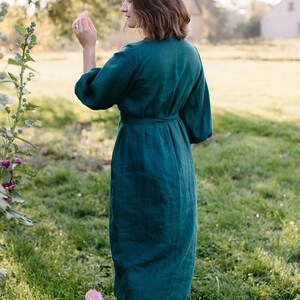 May include: A dark teal linen dress with a matching belt, three-quarter sleeves, and a knee-length hem. The dress is worn outdoors in a grassy area with pink flowers and a blurred background.