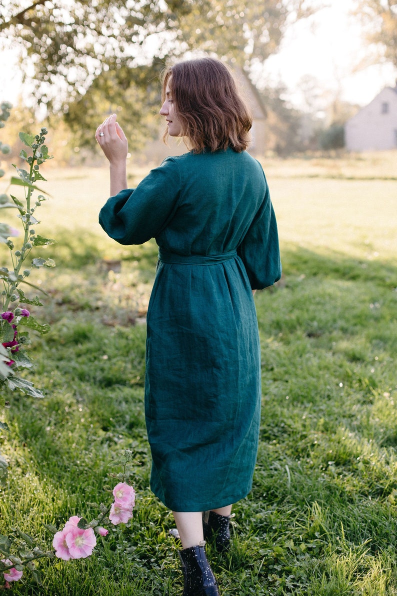 May include: A dark teal linen dress with a matching belt, three-quarter sleeves, and a knee-length hem. The dress is worn outdoors in a grassy area with pink flowers and a blurred background.
