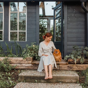 May include: A woman in a light-colored wrap dress sits on the steps of a dark gray shed with a small, fluffy, orange dog. The shed has a glass roof and windows, with plants visible inside and out.