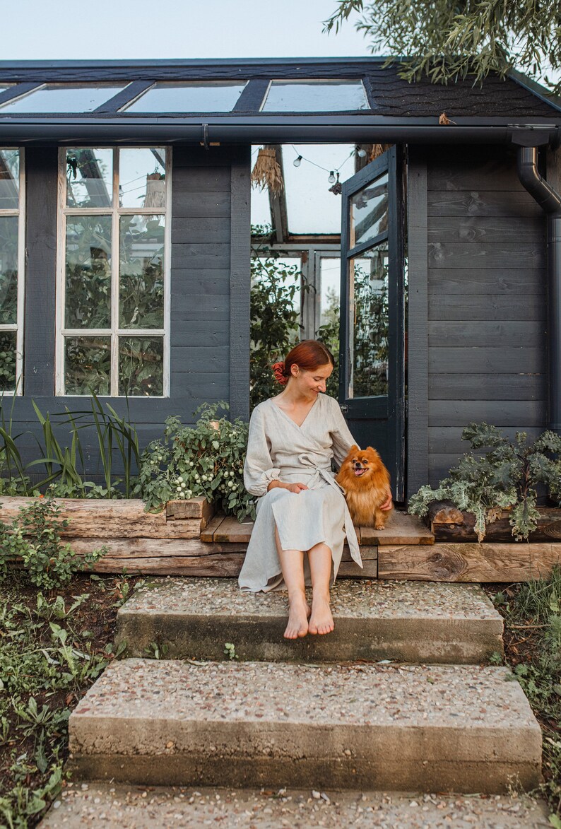 May include: A woman in a light-colored wrap dress sits on the steps of a dark gray shed with a small, fluffy, orange dog. The shed has a glass roof and windows, with plants visible inside and out.