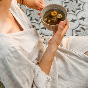 May include: A person holding a beige ceramic bowl filled with tea and flowers. The tea is a light amber color, and the flowers are yellow and white. The person is wearing a light beige linen robe.