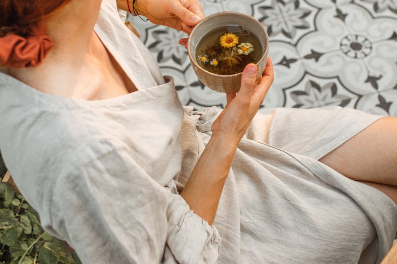 May include: A person holding a beige ceramic bowl filled with tea and flowers. The tea is a light amber color, and the flowers are yellow and white. The person is wearing a light beige linen robe.