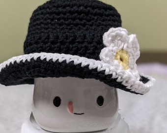 May include: A white ceramic mug with a hand-painted face wearing a black crocheted hat with a white crocheted flower.