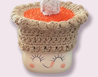 May include: A crocheted pumpkin pie mug cozy with a white pom pom on top. The cozy is beige with an orange pie crust and a white filling.