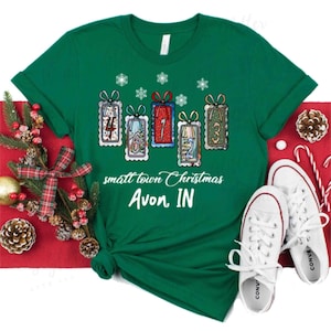 May include: A green t-shirt with a Christmas-themed design. The shirt features gift box illustrations with numbers, snowflakes, and the text "small town Christmas Avon, IN." White sneakers and holiday decorations are also in the image.