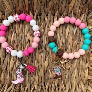 May include: Two beaded wrist keychains. One is pink, white, and red with a cowboy boot charm and a pink tassel. The other is pink, brown, and turquoise with a cowboy boot charm. Both are on a woven background.
