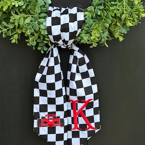 May include: A black and white checkered fabric decoration with a red race car and the letter "K" embroidered on it. The decoration is hanging from a green wreath. The race car has the number "96" on it.