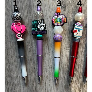 May include: Four decorated pens with different themes. The first pen has a black and white chevron pattern, a pink stethoscope, and the text "Have the day you deserve". The second pen has a purple body with a skeleton hand and the text "Have the day you deserve". The third pen has a white body with an orange apple and the text "Teacher". The fourth pen has a red body with a cartoon bottle and the text "Ultrasound Gel".